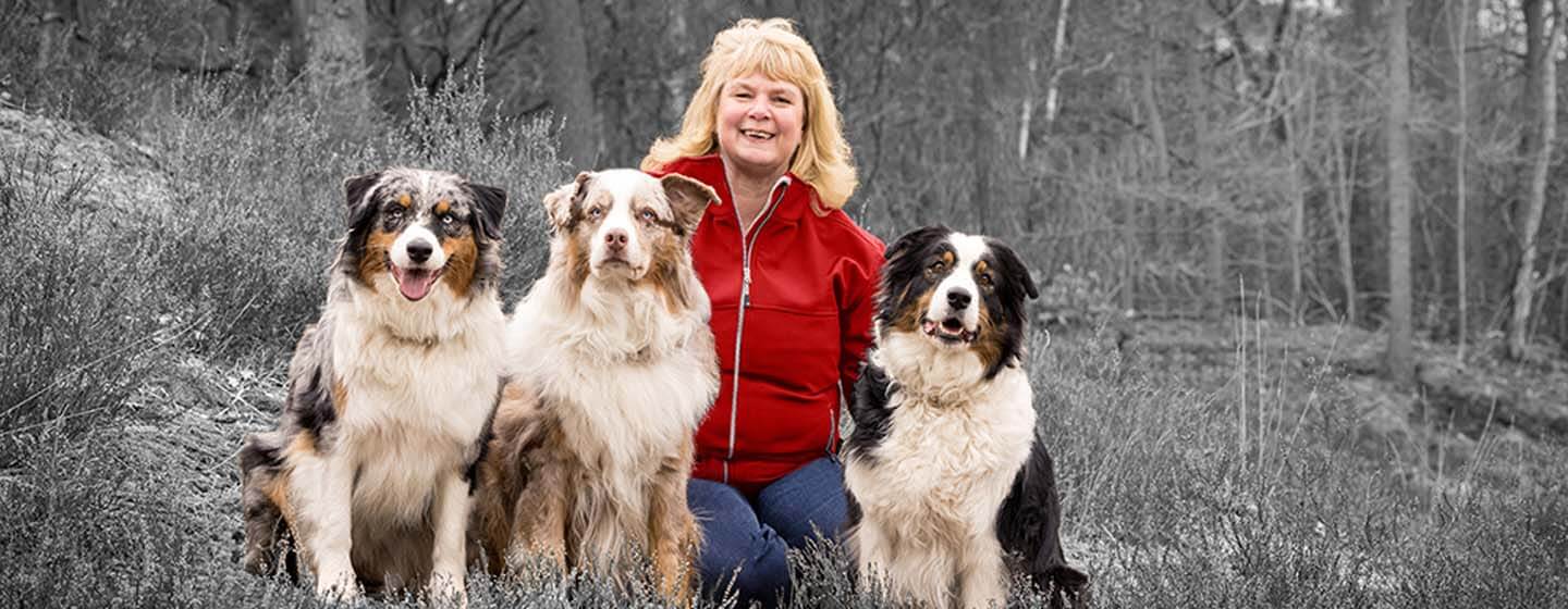 Smiling woman with three dogs in an outdoor setting, highlighted by selective color.